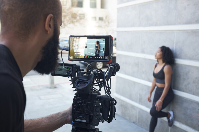 Videographer operating a cinema camera with external monitor on set while filming a female subject against a concrete wall.