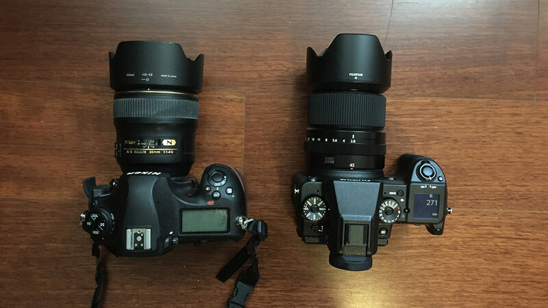 Two DSLR cameras with telephoto lenses and lens hoods positioned side by side on a wooden surface.
