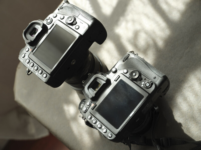 Two professional DSLR cameras positioned side by side on a light surface, showing rear LCD screens and control buttons.
