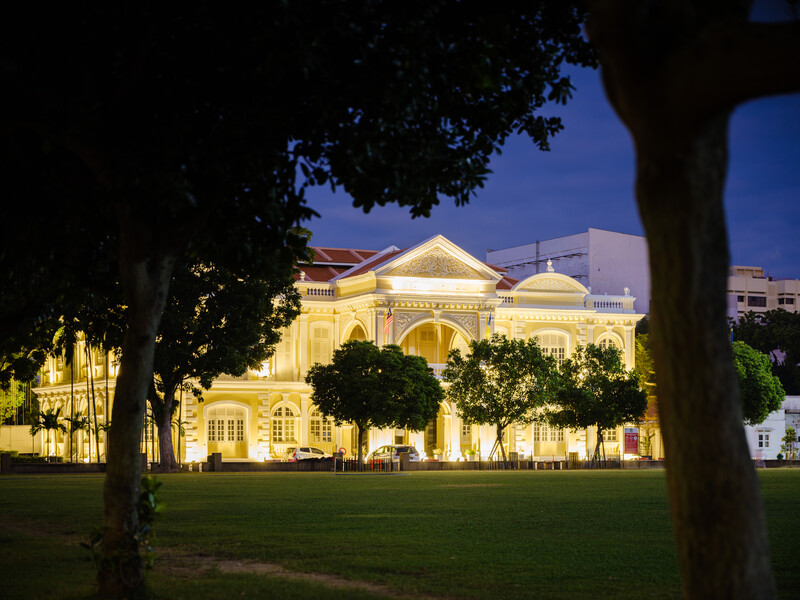 Neoclassical building illuminated at dusk with warm lighting, framed by silhouetted trees.