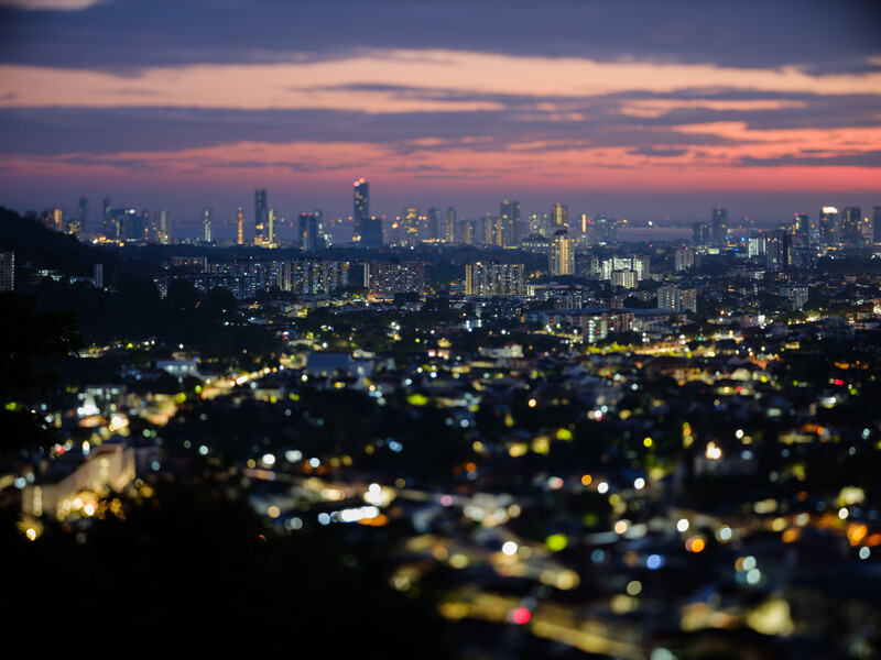 Cityscape at twilight with illuminated neighborhoods in foreground and distant skyline silhouetted against purple sunset sky.