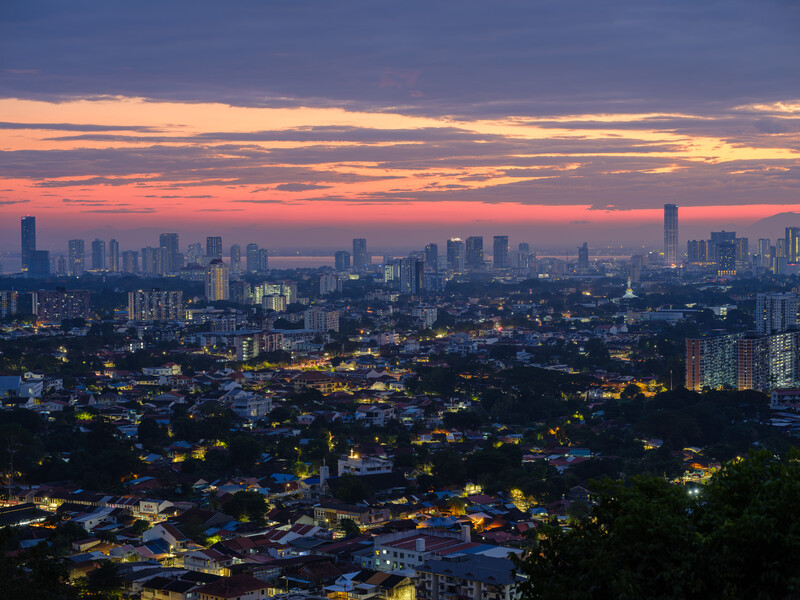 Twilight cityscape with illuminated buildings in foreground and downtown skyline silhouetted against purple and orange sunset sky.