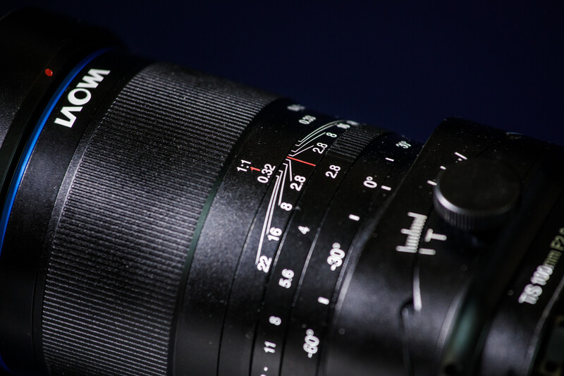 Close-up of a manual focus lens showing aperture ring, focus ring, and distance scale markings against dark background.