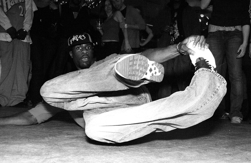 Dancer performing a floor move with extended legs while surrounded by spectators in low-light indoor setting.