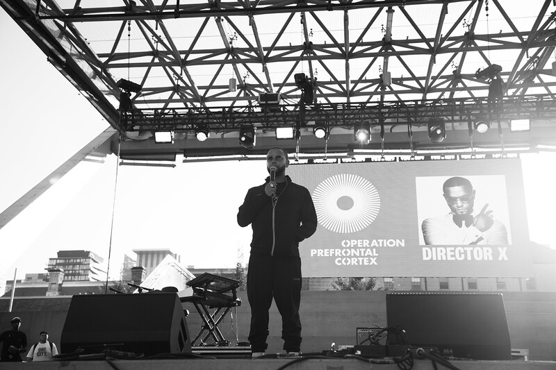 Speaker at outdoor event stage with professional lighting rig and projection screens displaying text and portraits.