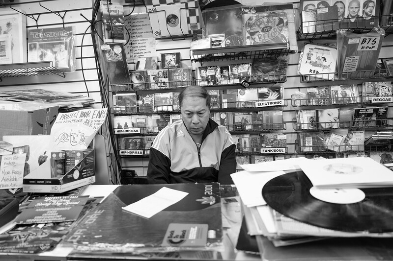 Man seated at a cluttered desk surrounded by photographs, posters, and memorabilia pinned to walls behind him.