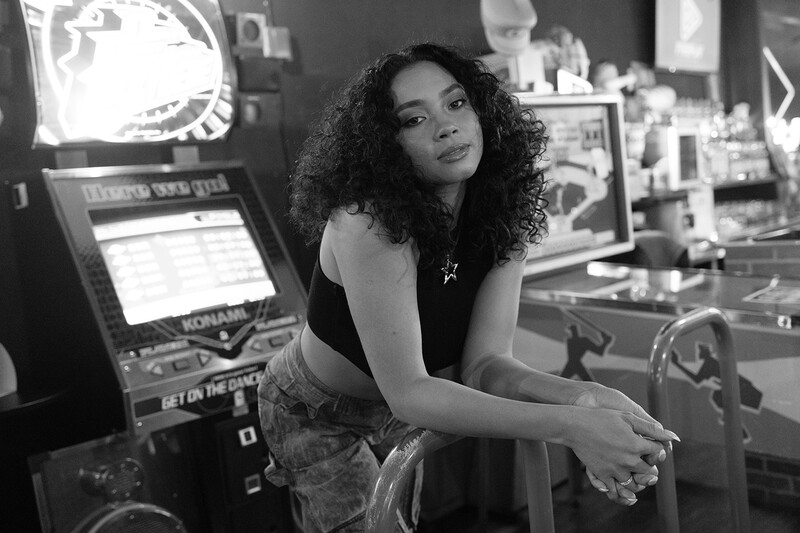 Woman with curly hair in arcade setting, leaning against gaming machine in black and white photography.