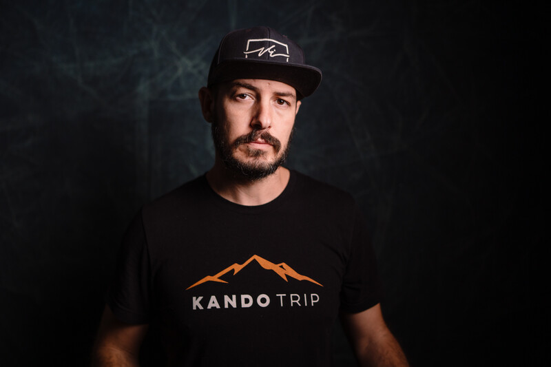 Man wearing black cap and KandoTrip branded t-shirt against dark textured background.
