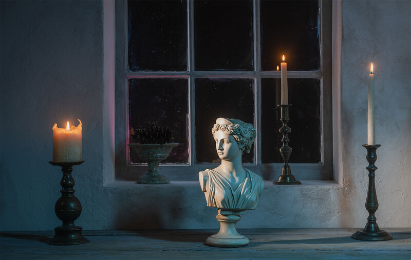 Classical bust sculpture centered between candlesticks on a windowsill at dusk, with a potted plant visible through the window behind it.