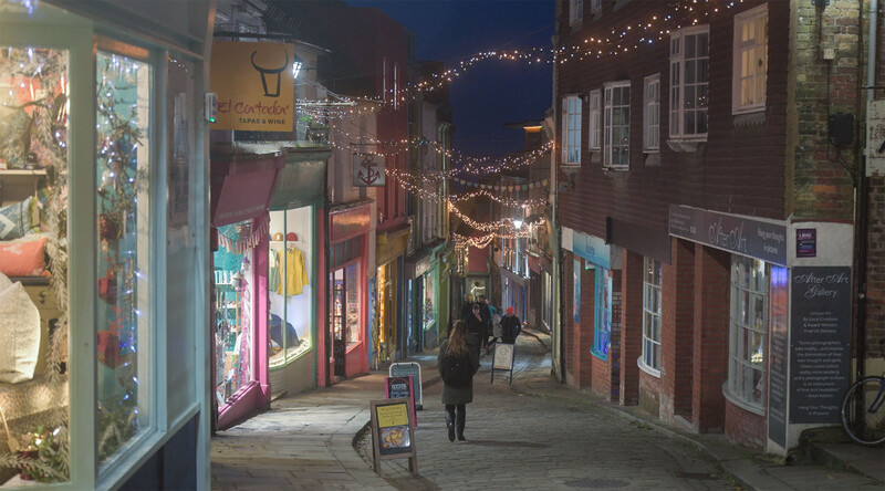 Illuminated storefront row along a narrow evening street decorated with warm string lights overhead.