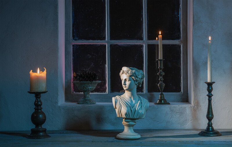 Classical bust sculpture displayed on a windowsill between lit candles in candlesticks against a blue wall.