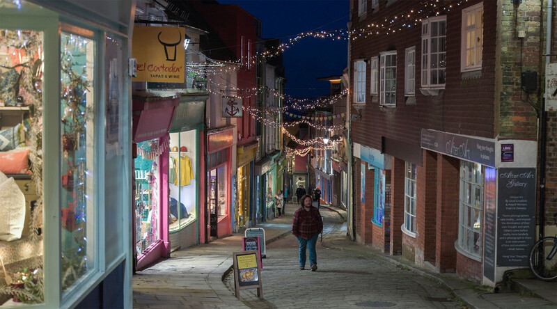 Narrow European street at dusk lined with illuminated storefronts and decorative lights overhead.