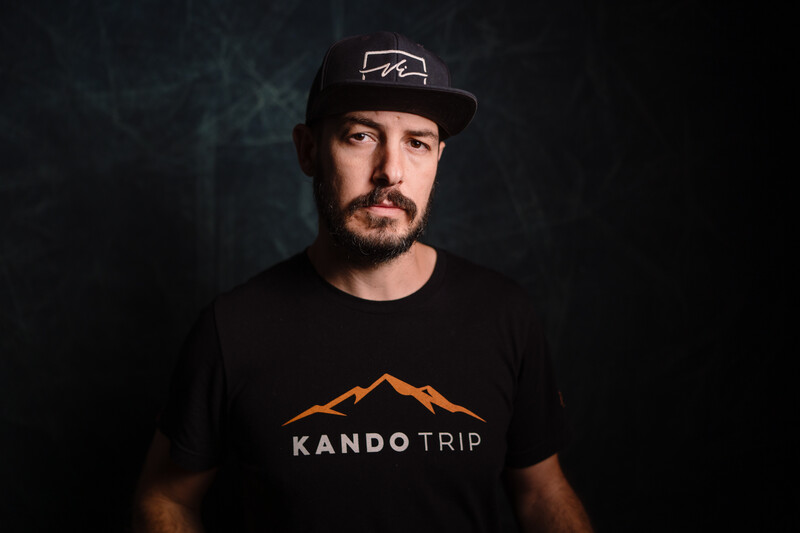 Man wearing black cap and Kando Trip branded t-shirt against dark background.