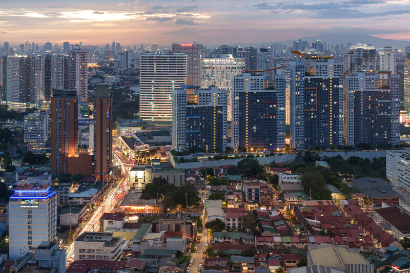 Urban cityscape at dusk with illuminated high-rise buildings, traditional architecture in foreground, and snow-capped mountain in distant background.