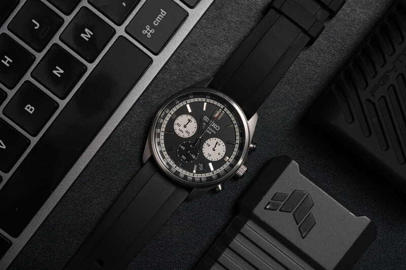 Chronograph watch with black dial and white subdials on black leather strap, photographed flat lay with keyboard and desk accessories.