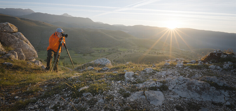 Photographer in orange jacket using tripod to capture landscape at golden hour from rocky hilltop.