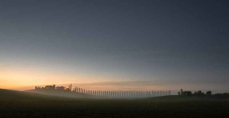 Tuscan villa silhouetted against golden sunrise with cypress tree lined road emerging from morning fog.