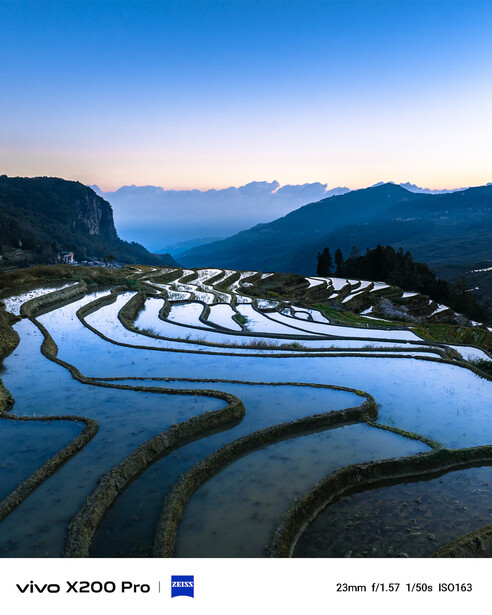 Terraced rice paddies flooded with water reflecting the sky at dawn, with mountains silhouetted in the background.