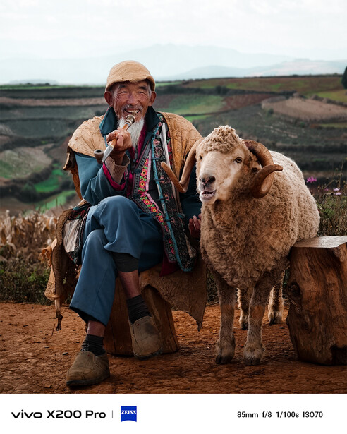 Portrait of a person crouching beside a sheep in a rural landscape with terraced fields in the background.
