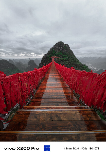 Long wooden bridge with bright red fabric railings leading toward a steep green mountain peak under overcast sky.