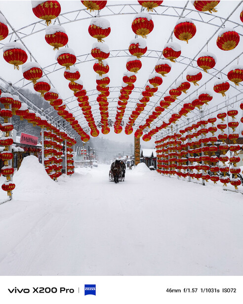 Red lanterns suspended symmetrically above a snow-covered street with buildings and a lone figure below.