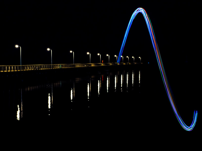 Nighttime photograph of an illuminated pier with blue neon light trails forming a curved arch at the end.