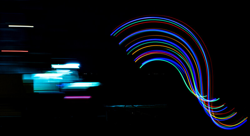 Light painting photograph with cyan geometric shapes on the left and multicolored curved light trails on the right against a black background.