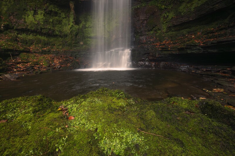 Waterfall cascading into a dark pool surrounded by moss-covered rocks and forest vegetation.