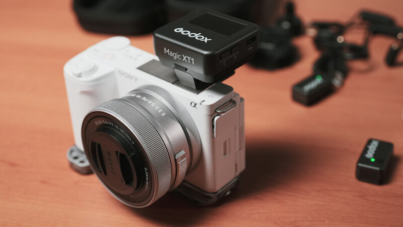 White mirrorless camera with silver lens mounted on orange surface, with black wireless flash unit attached on top.