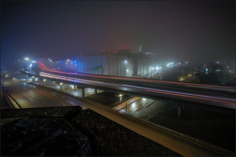 Long exposure photograph of a highway overpass at night with light trails from vehicle headlights and taillights streaking across the frame.