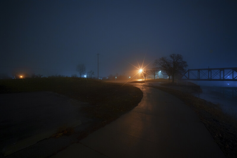 Rural landscape at dusk with wet ground, distant street lights, and a bridge silhouetted against twilight sky.