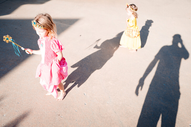Two young children in colorful dress-up clothes standing on pavement with adult shadows visible.