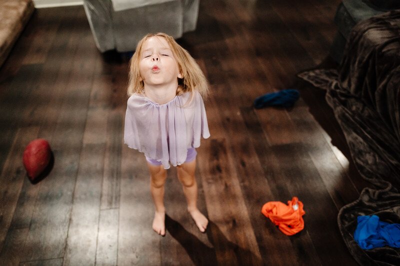 Young child in lavender dress standing on wooden floor surrounded by scattered clothing.