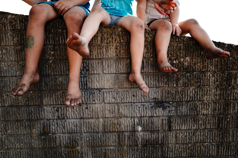 Four children sitting on a textured brick wall, legs dangling, wearing casual summer clothing.