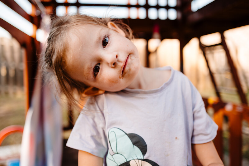 Toddler in light blue t-shirt tilting head and looking at camera with curious expression.