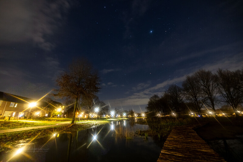 Riverside park at twilight with warm street lamps reflecting in still water, bare trees lining both banks, and stars visible in the darkening sky.
