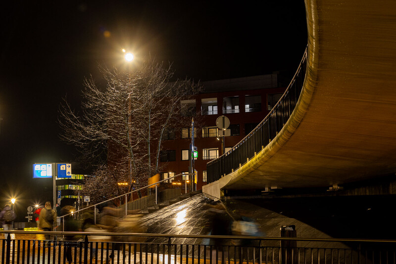 Modern curved pedestrian bridge illuminated at night over dark water with lit building and bare tree.