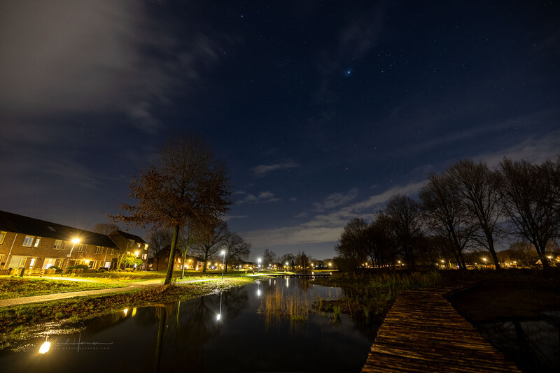 Nighttime canal scene with illuminated buildings, wooden dock, and bare trees reflected in still water.