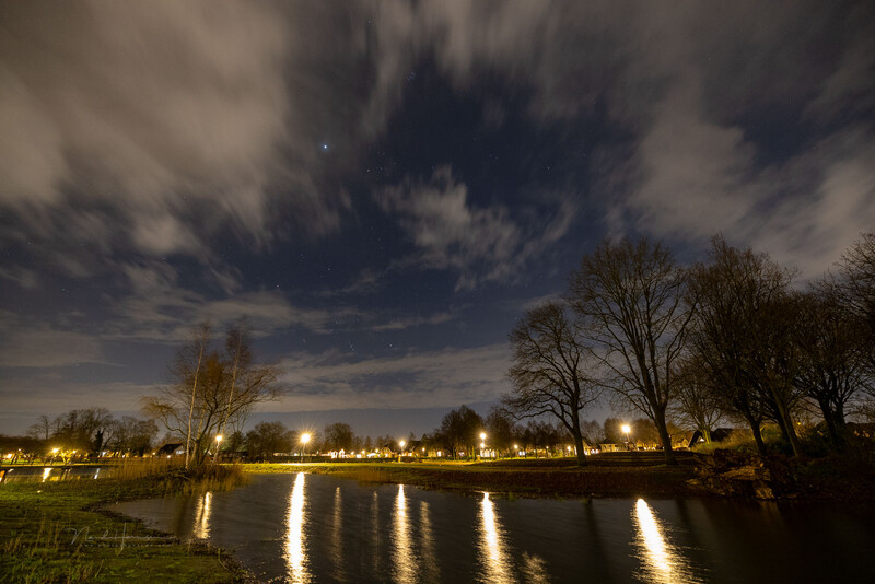 Nighttime landscape with bare trees silhouetted against dramatic cloudy sky, reflecting in still water below with streetlights along the distant bank.