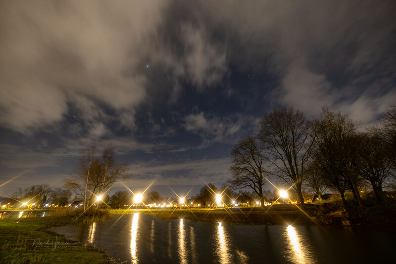Nighttime landscape with bare trees silhouetted against dramatic clouds and street lights reflecting in still water.