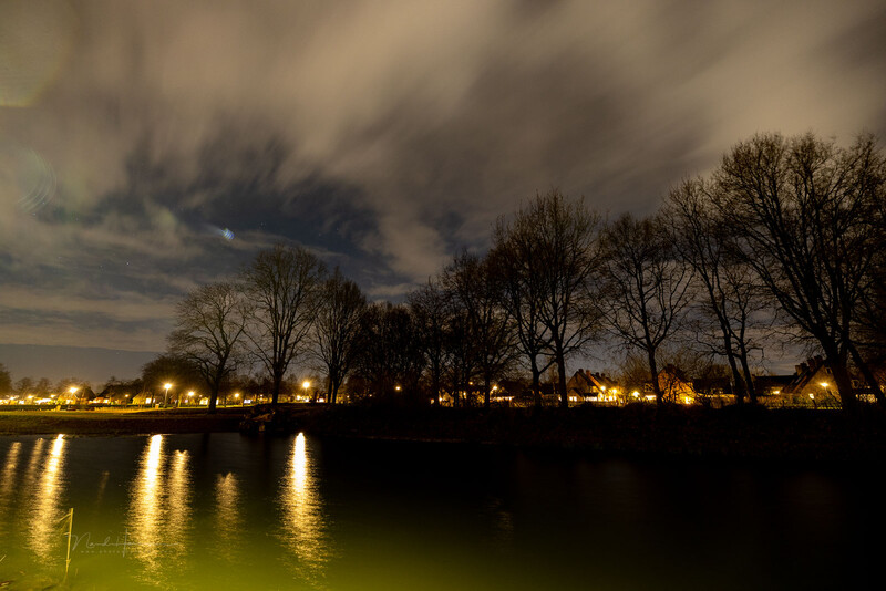 Bare trees silhouetted against a dramatic cloudy sky, reflected in still water lit by warm streetlights along a riverbank at dusk.