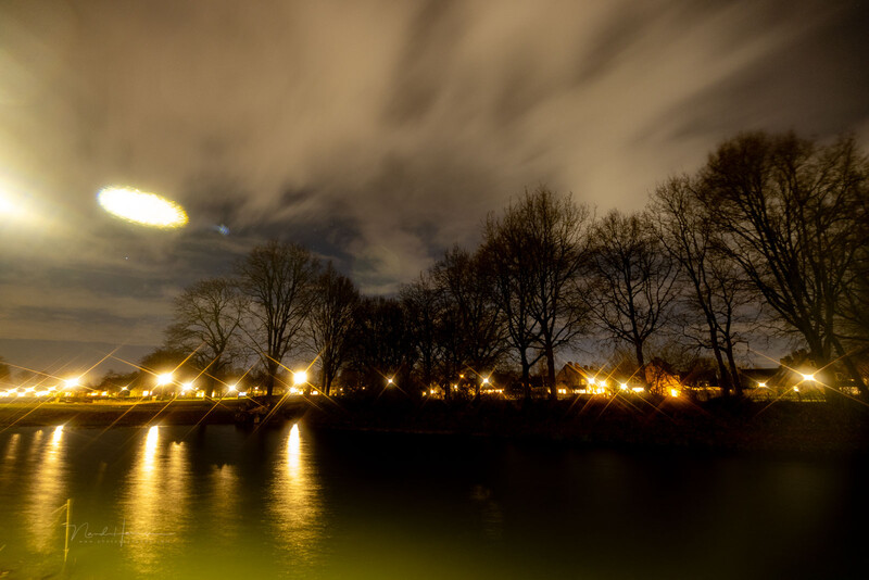 Nighttime waterside scene with bare trees silhouetted against a cloudy sky, their reflections mirrored in still water below street lamps.