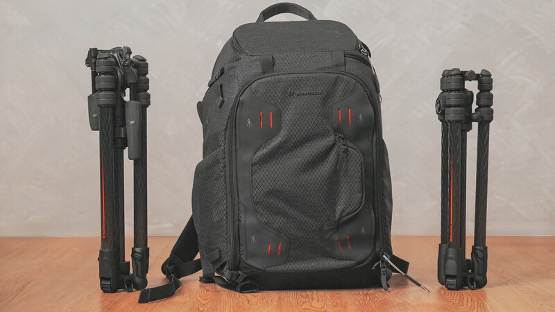 Black camera backpack with red accents displayed between two compact tripods on wooden surface.