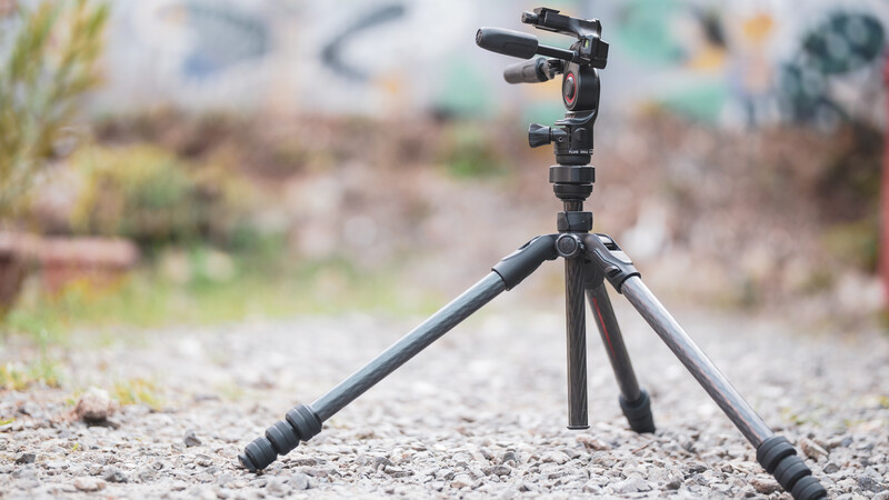 Compact tripod with ball head and microphone mount on gravel ground with blurred background.