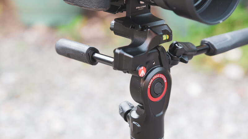 Close-up of a camera gimbal head with red accents mounted on a tripod, showing the control dial and pan handles.