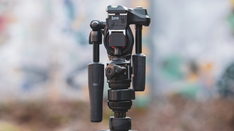 Three-axis motorized gimbal with smartphone mounting head and extended handle grips against blurred background.