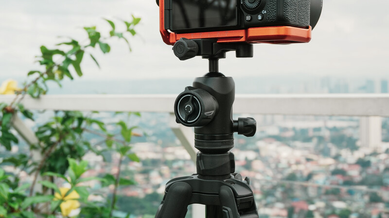 Orange and black mirrorless camera mounted on tripod with ball head, overlooking urban cityscape and water.