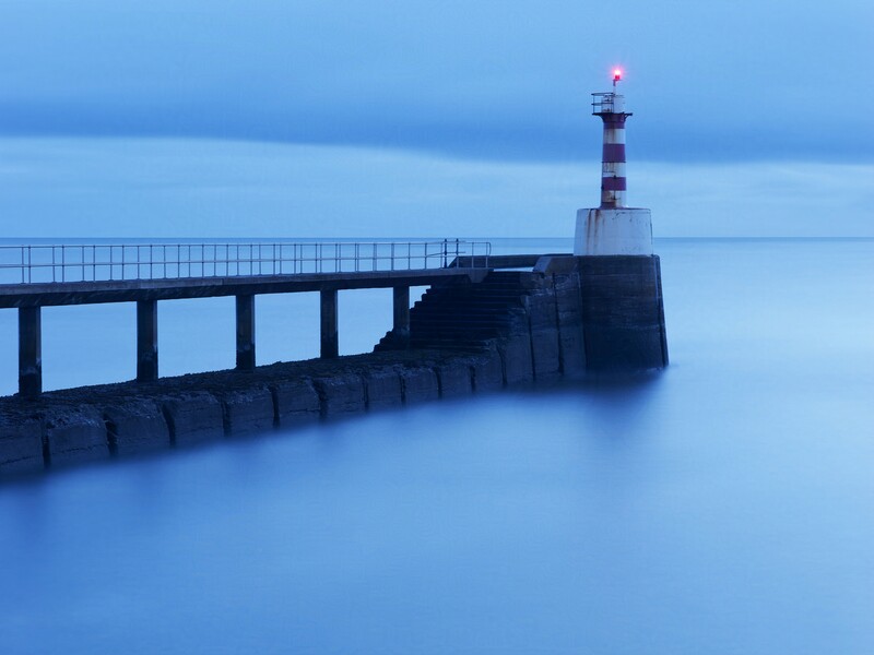 Lighthouse on a stone pier extending into calm blue water at dusk with a long-exposure effect.