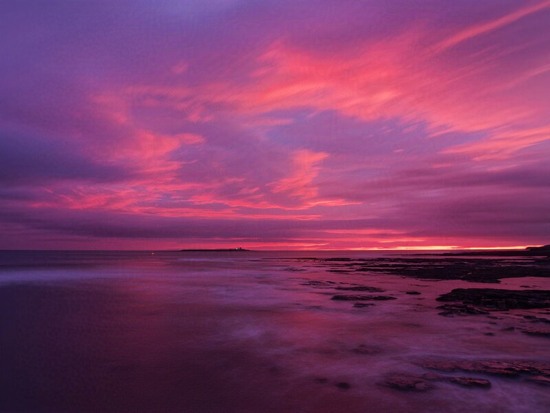 Dramatic purple and pink sunset over a rocky coastline with still water reflecting the sky.
