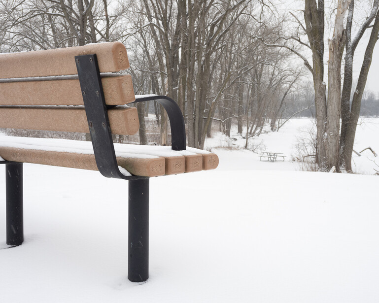 Wooden park bench with black metal frame covered in snow, bare trees visible in background.